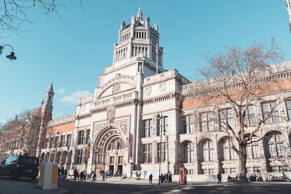 A wide-angle view of a historic stone building with an elaborate central clock tower and intricate architectural details, situated on a busy street with pedestrians walking along the pavement. Several leafless trees line the sidewalk, and a pavement parking area is present with a few vehicles, including a black van, parked nearby. The building’s entrance is framed by arched doorways and decorative stone carvings. The sky above is clear and blue, and the scene captures the typical street environment in central London, relevant to house removals and moving logistics in the vicinity of Tate Britain, as featured on removalspimlico.com.