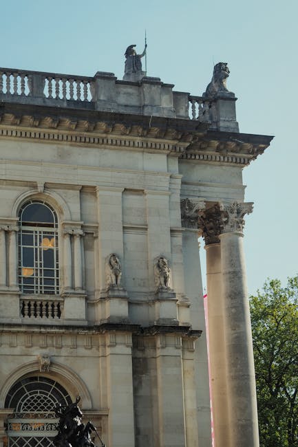 Image of the upper portion of a classical-style building facade featuring decorative architectural elements such as columns, ornate moldings, and sculptures of seated figures along the balustrade on the roofline. The building is constructed from light-colored stone or concrete, with large arched windows visible on the lower level. The sky is clear, and some green trees are seen to the right, indicating an outdoor setting. This architectural detail aligns with the surroundings near Tate Britain in Pimlico, where [COMPANY_NAME] offers professional house removals and furniture transport services, including packing and loading processes for home relocation projects.